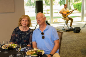 Two people seated at a banquet table smiling for the camera with a guitar player in the background