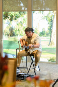 Man playing a guitar in front of a window overlooking a golf course
