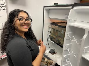 smiling student standing by a small classroom aquarium in a mini refrigerator