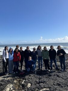 A group of twelve people standing and posing on a rocky beach with waves in the background under a clear blue sky. Some are wearing jackets and hoodies. One person is kneeling in front.