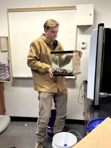 Person in a brown jacket holds a small aquarium with rocks and a turtle inside. They stand in an office or classroom with a whiteboard behind them, near a desk and a blue bucket.