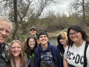 A group of six people standing outdoors near a creek, smiling at the camera. They are surrounded by trees and wearing casual clothing. The sky is cloudy.
