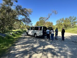 A group of people stands near a white van parked on a sunny dirt road, surrounded by trees and greenery under a clear blue sky.