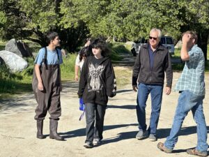 Five people walk together outdoors on a sunny day, surrounded by trees. One person wears fishing waders, another wears a black hoodie with a design, and the others are in casual clothes, chatting as they walk on a dirt path.