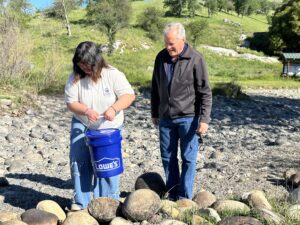 A young woman holding a blue Lowe’s bucket stands on rocky ground near a stream, while an older man in a dark jacket stands beside her. There is greenery and hills in the background on a sunny day.