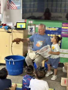 An older man uses a brown dog puppet to entertain a group of young children sitting on the floor. A smiling boy holds a white laundry basket next to him. Classroom decorations and a TV are visible in the background.