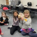 A group of three children sitting on a classroom floor. Two girls in the middle are holding a large seashell together and smiling. Chairs and tables are arranged against the wall in the background.