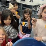 A group of six children are gathered around a blue container, smiling and holding seashells. They're wearing casual clothes, and some have orange stickers on their shirts. The setting appears to be a classroom with desks and chairs in the background.