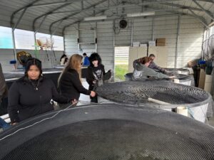 Several people stand around large circular tanks inside a metal building, examining the tanks and interacting with each other. The space appears to be a research facility or aquaculture lab.