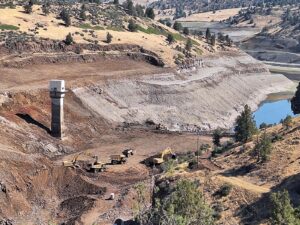 A drained reservoir with exposed earth slopes, construction vehicles, and a tall concrete intake tower; surrounding hills are covered with sparse vegetation and patches of trees.